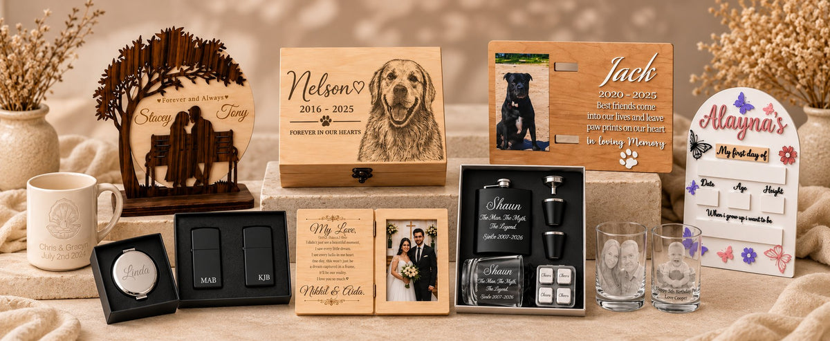 A curated gift set display featuring personalized wooden keepsake boxes, engraved photo plaques, a wooden bench silhouette ornament, engraved glass bottles, two clear drinking glasses, and small decorative items arranged on a beige fabric surface with dried floral accents.